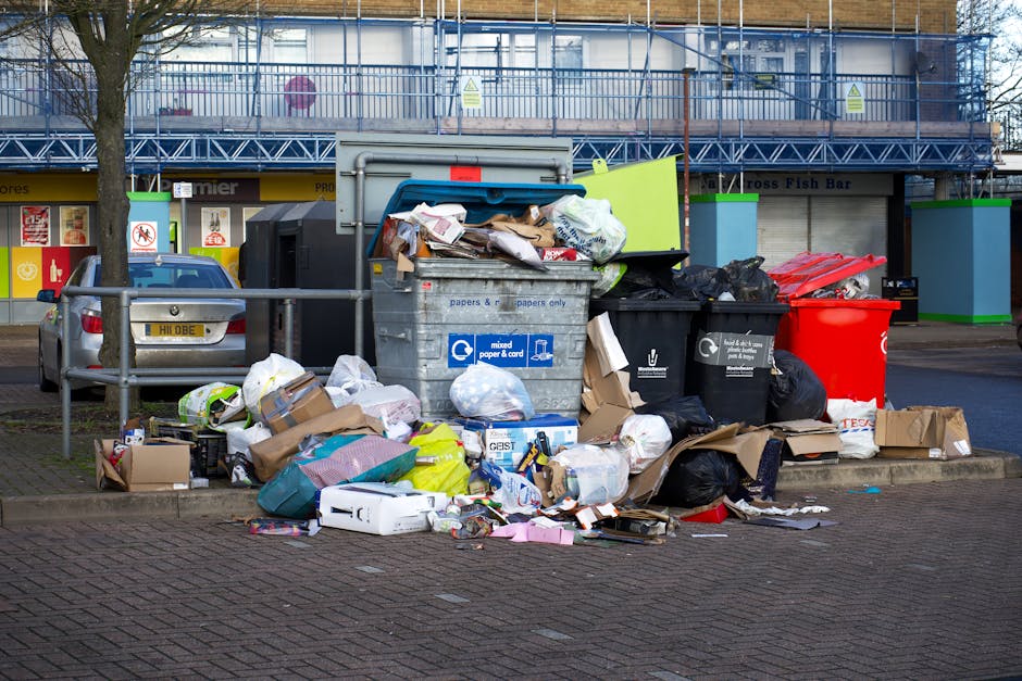 A collection of overflowing rubbish bins and scattered waste on a paved sidewalk, situated near a metal railing and a parked grey car with a yellow license plate. The waste includes a large grey mixed-paper-and-cardboard recycling bin with its lid open, overflowing with crumpled paper, cardboard boxes, and plastic wrappers; a black general waste bin with a partially open lid, filled with black plastic bags and other refuse; and a bright red recycling bin positioned next to the black bin. Surrounding the bins, there are numerous loose cardboard boxes, paper bags, and plastic packaging items, some flattened or torn. The background features a building with scaffolding, suggesting ongoing construction or renovation, along with storefronts and signage visible on the ground level. The scene appears to be in an urban area and illustrates the kind of waste accumulation that might occur when private rubbish collection services, such as those offered by Waste Removal Shoreditch, intervene in waste management and clearance activities in a busy town setting, emphasizing the importance of proper rubbish disposal and alternative waste handling methods.