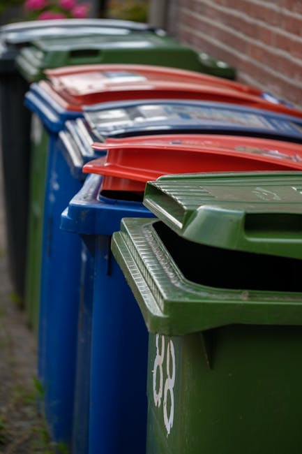 A row of public trash bins positioned along a pavement, with the nearest bin being green and featuring a sloped, ribbed lid. Behind it, several other bins are visible, including blue, red, and dark green, each with similar lids designed for rubbish disposal. The bins are aligned tightly next to a brick wall, suggesting an urban or residential setting. The green bin in the foreground has a slightly reflective surface, with subtle signs of weathering and dirt on the lid structure. The background includes part of the pavement and a blurred section of the brick wall, with ambient natural light illuminating the scene. This arrangement of multiple waste containers exemplifies common arrangements for communal or private waste collection, possibly used as an alternative to council-led rubbish collection services, supporting the context of independent disposal or on-site clearance services provided by Waste Removal Shoreditch.