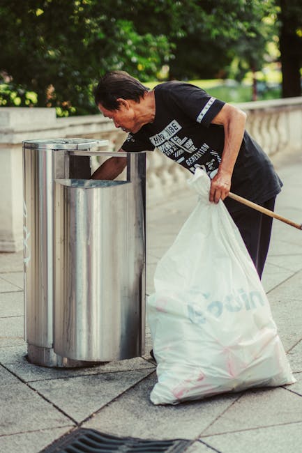 A collection of overflowing rubbish bins and scattered waste on a paved sidewalk, situated near a metal railing and a parked grey car with a yellow license plate. The waste includes a large grey mixed-paper-and-cardboard recycling bin with its lid open, overflowing with crumpled paper, cardboard boxes, and plastic wrappers; a black general waste bin with a partially open lid, filled with black plastic bags and other refuse; and a bright red recycling bin positioned next to the black bin. Surrounding the bins, there are numerous loose cardboard boxes, paper bags, and plastic packaging items, some flattened or torn. The background features a building with scaffolding, suggesting ongoing construction or renovation, along with storefronts and signage visible on the ground level. The scene appears to be in an urban area and illustrates the kind of waste accumulation that might occur when private rubbish collection services, such as those offered by Waste Removal Shoreditch, intervene in waste management and clearance activities in a busy town setting, emphasizing the importance of proper rubbish disposal and alternative waste handling methods.
