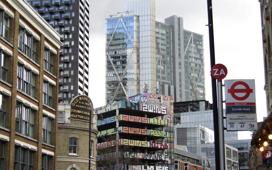 A street scene in an urban area featuring a tall, modern glass skyscraper in the background, with reflective windows showing the surrounding cityscape. In the foreground, there are older brick buildings with multiple rectangular windows, one with white framed sashes. Prominently visible are several street signs, including a red bus stop sign indicating a route towards Shoreditch Church, and a round bus stop marker on a pole. Partially obstructing the view is a colorful billboard with multiple layered, stacked panels displaying graffiti-style text and patterns in bright colors such as green, orange, purple, and black. A smaller sign above mentions 'Old Blue A.D. 1971,' referencing a building or business in the vicinity. The surroundings are well-lit with natural daylight, capturing a lively city atmosphere associated with public transit and urban commerce.
