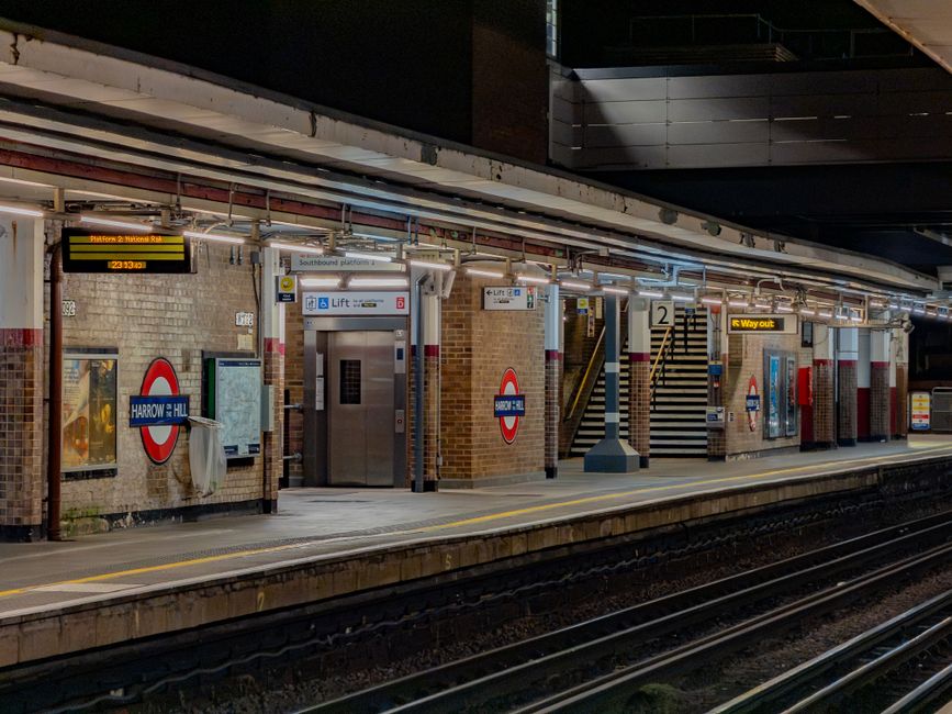A nighttime view of Harrow & Wealdstone underground station platform showing brick and tiled walls with signage indicating the Harrow & Wealdstone line. There are two elevators with metallic doors and clear signage, positioned centrally among the brickwork. To the right, a staircase with black and white striped safety markings leads up to the street level, with metal railings and illuminated signs overhead. The platform surface is paved with concrete, edged by railway tracks visible in the foreground, and lit by overhead lighting creating a well-lit environment typical of urban underground stations. The space appears clean and maintained, with clear visual indicators for passengers, including directional and safety signs. The scene is consistent with urban transportation infrastructure, encompassing elements of public transit and station amenities, suitable for a blog post related to public transport or transportation infrastructure management.